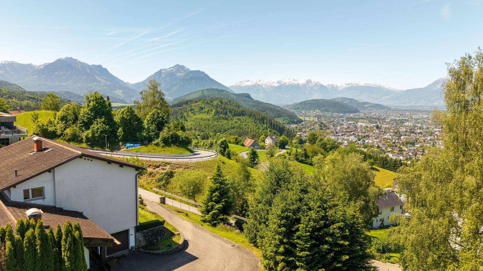 Weitläufiger Ausblick über die grüne Landschaft, eine Stadt und die beeindruckende Bergkette.