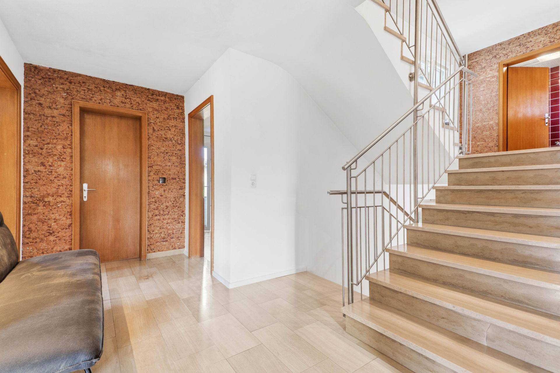 Heller Flur mit Holztreppe, Metallgeländer und gemusterten Tapeten an den Wänden.