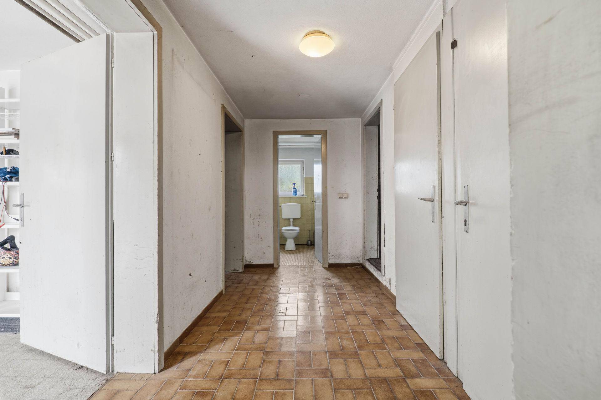Älterer Flur mit braunen Fliesen und Blick auf eine Toilette im Hintergrund.