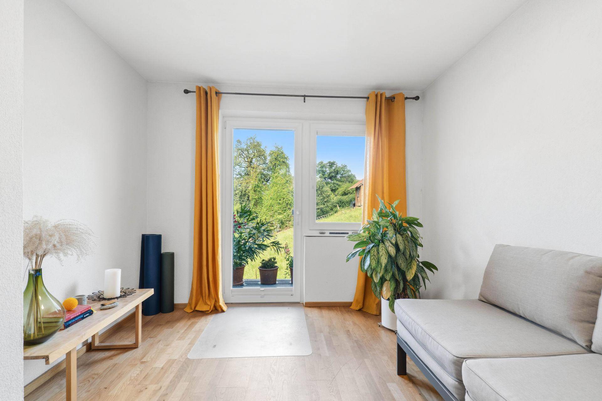 Helles Zimmer mit Holzboden, gelben Vorhängen und Zugang zu einem kleinen Gartenbereich.