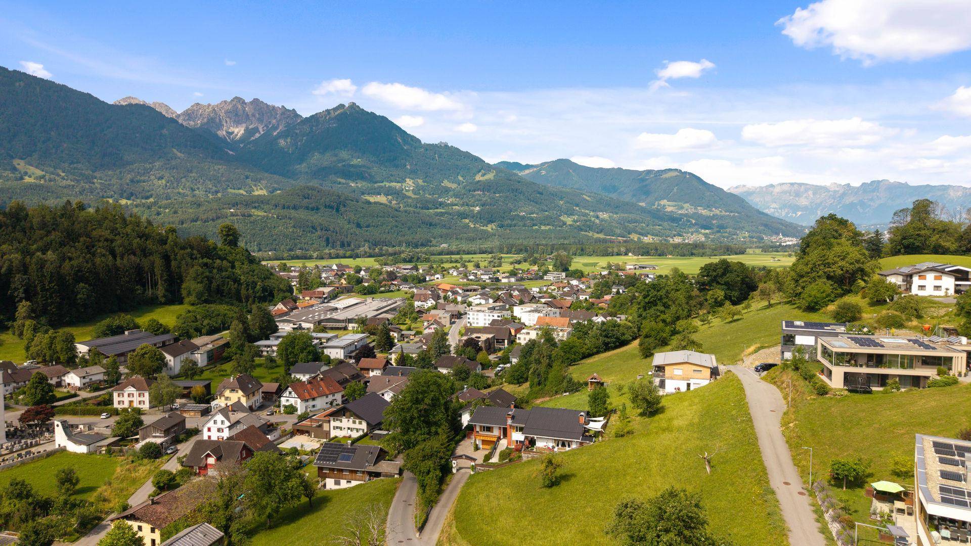 Panoramablick auf ein Dorf in einem Tal, umgeben von grünen Bergen und Wäldern.