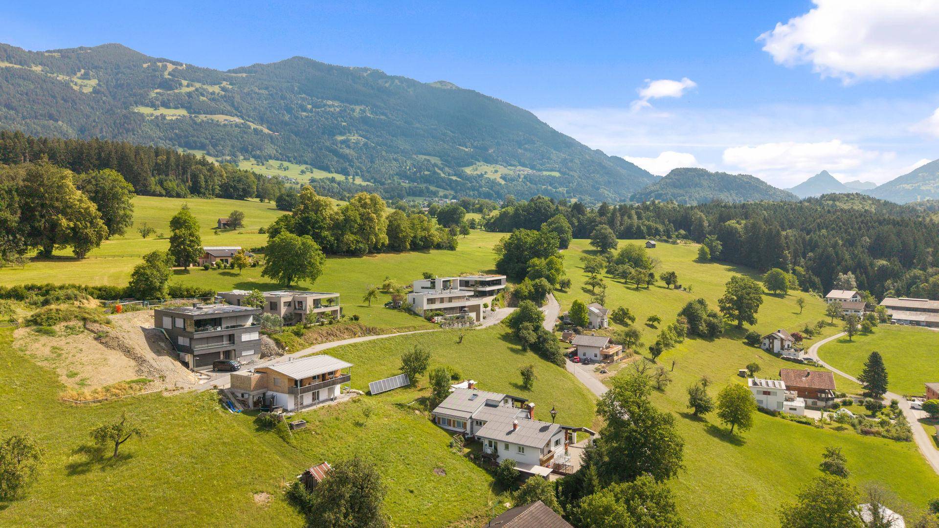 Weitläufige Luftaufnahme einer ländlichen Wohngegend mit grünen Hügeln und Bergen im Hintergrund.