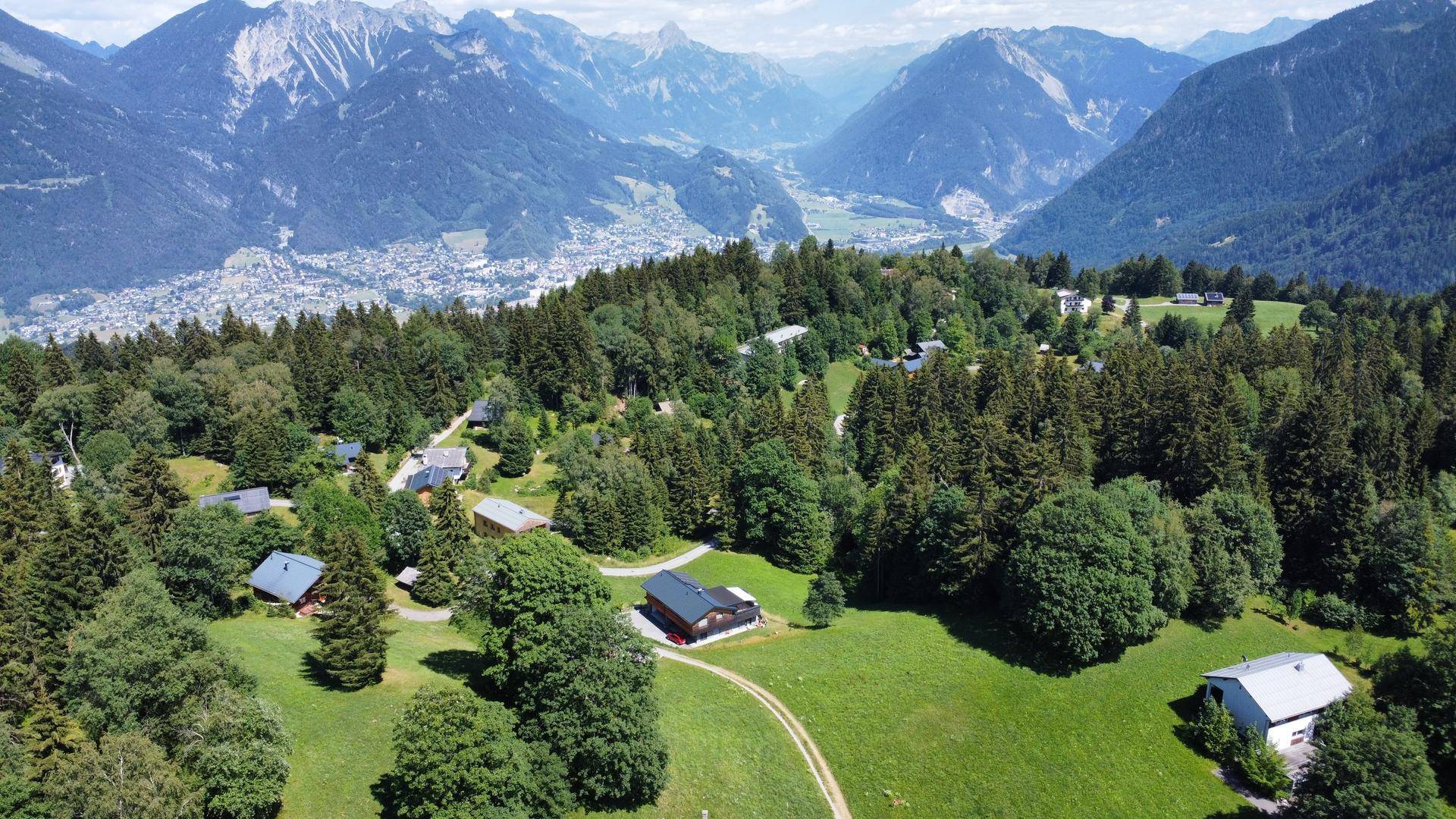 Weitläufige Luftaufnahme einer Siedlung in einer idyllischen Berglandschaft mit bewaldeten Hängen.