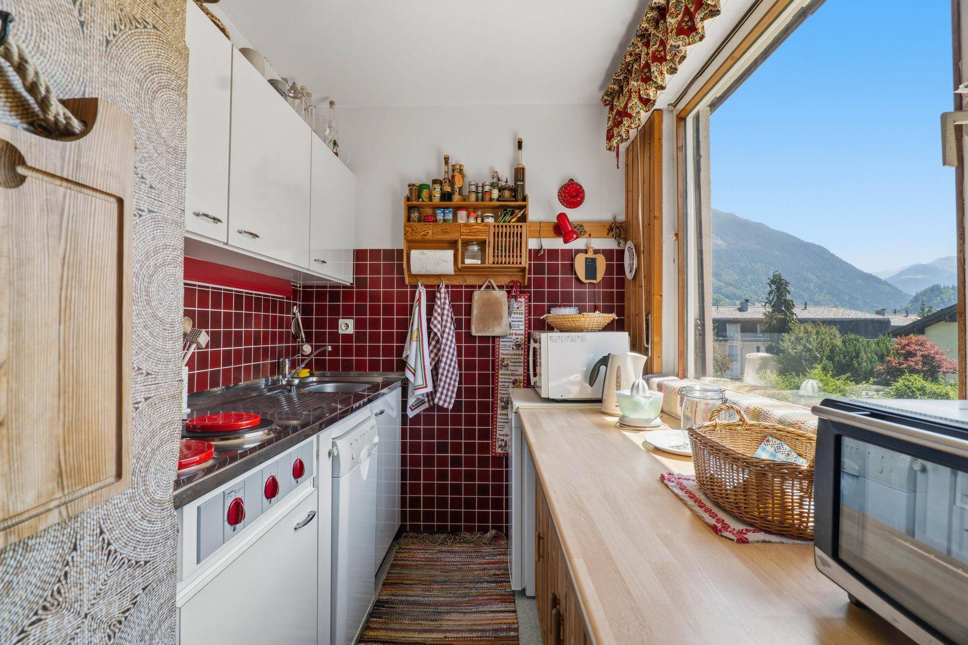 Kleine Küche mit roten Fliesen, weißen Schränken und einem Fenster mit Bergblick.
