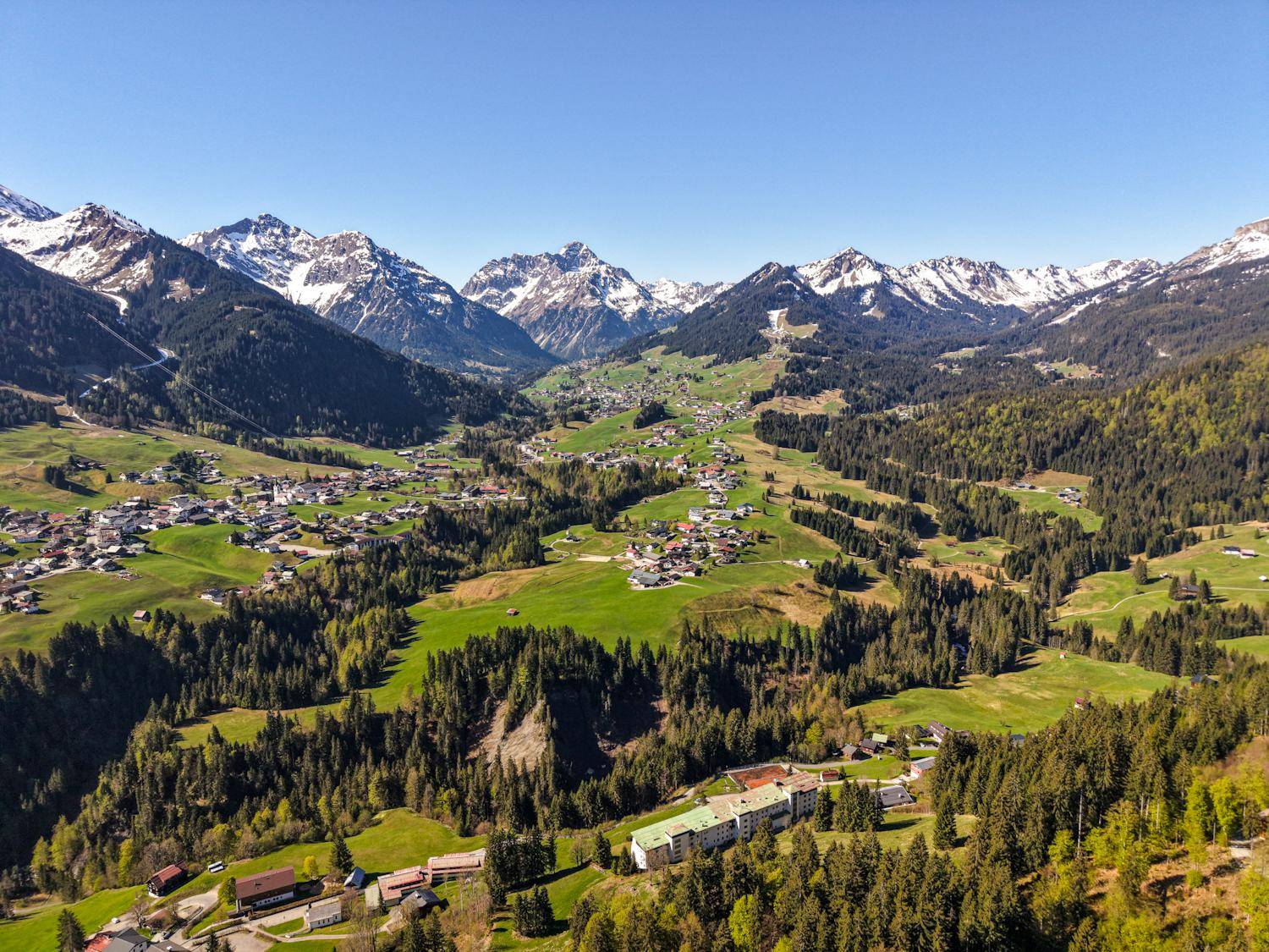 Luftaufnahme eines grünen Tals mit Dörfern und schneebedeckten Bergen.