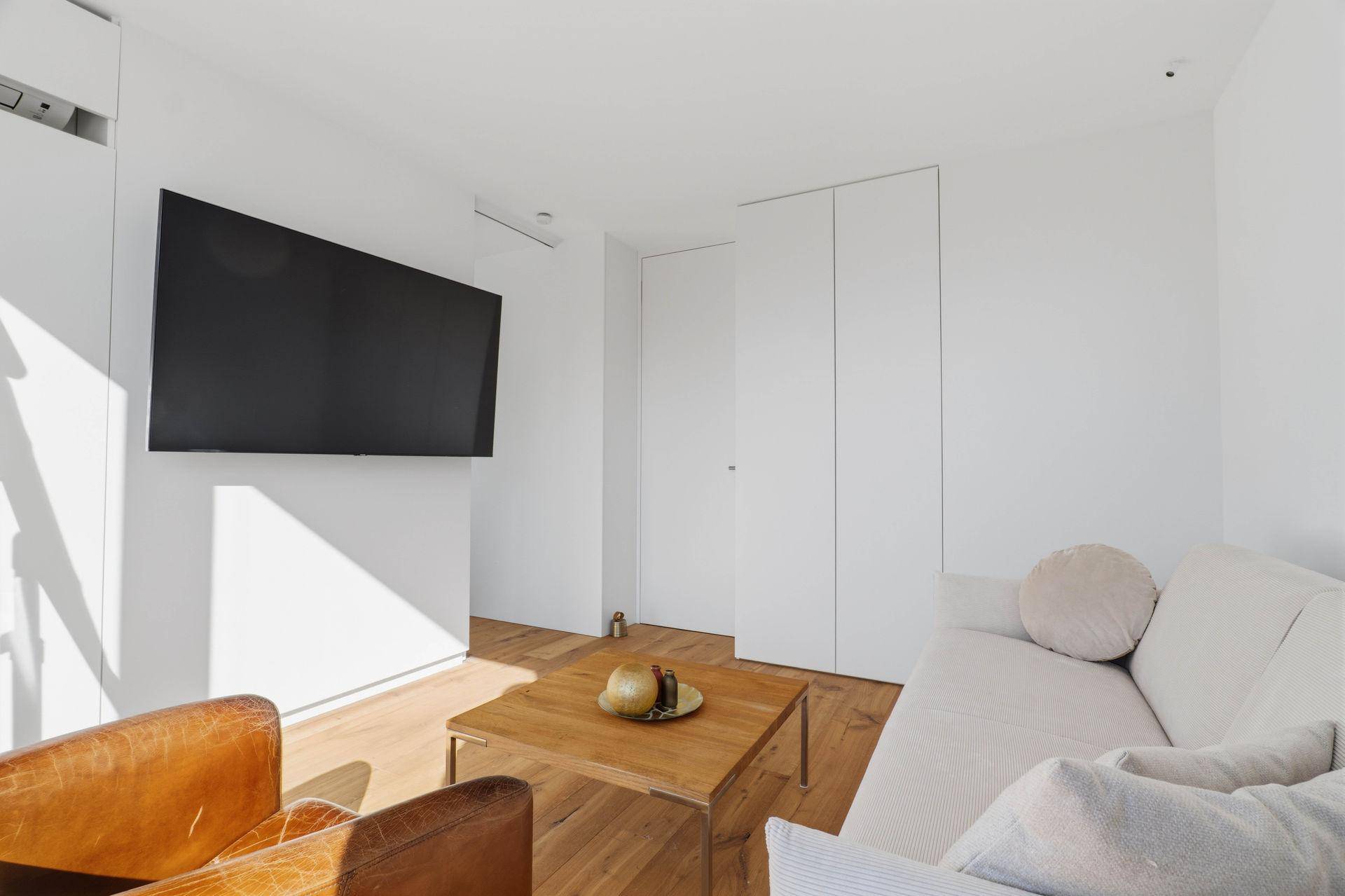 Heller Wohnbereich mit Wandfernseher, Sofa und integrierten weißen Schränken.