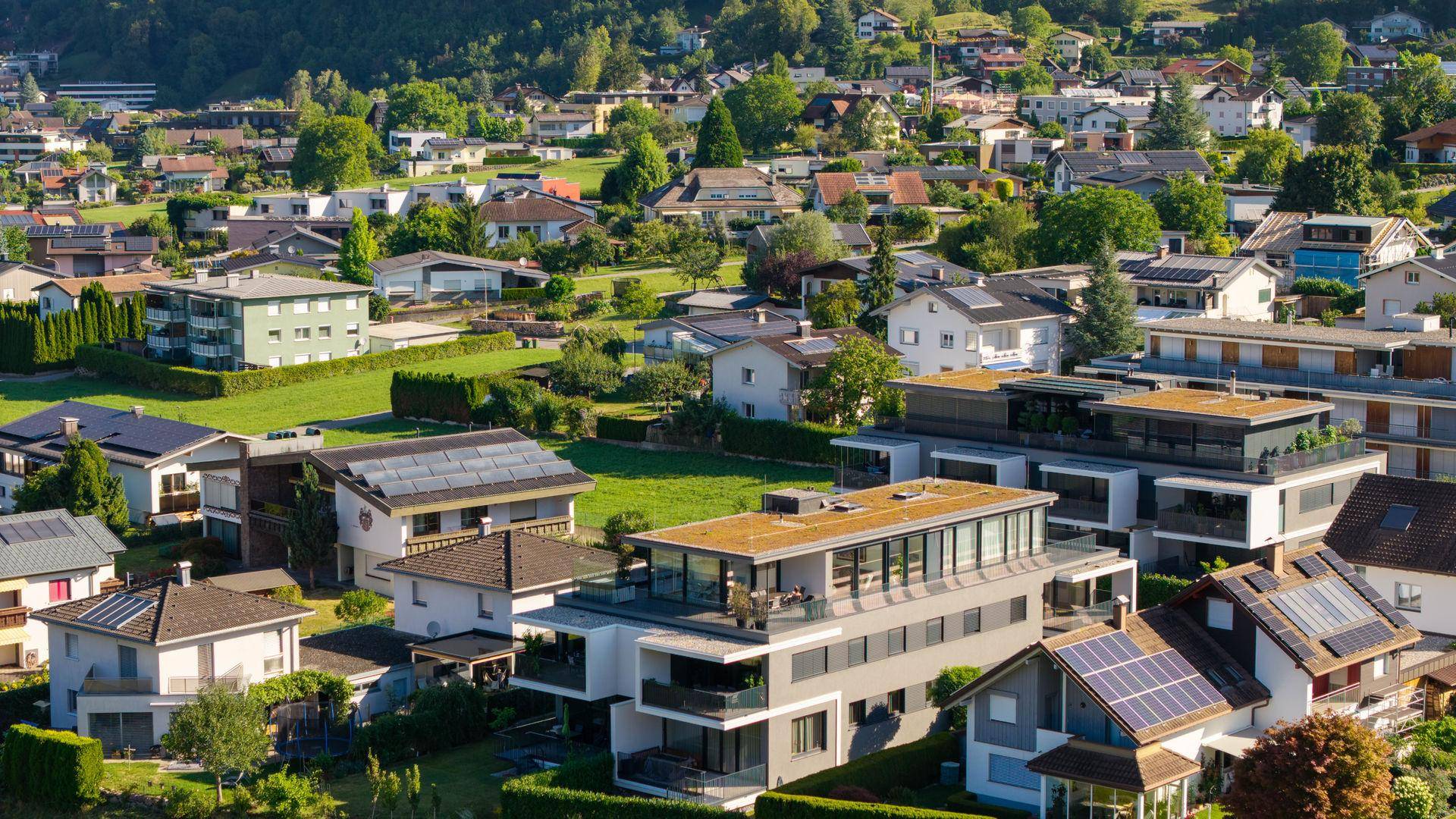 Luftaufnahme einer Wohnsiedlung mit modernen Gebäuden und grünen Flächen vor alpiner Kulisse.