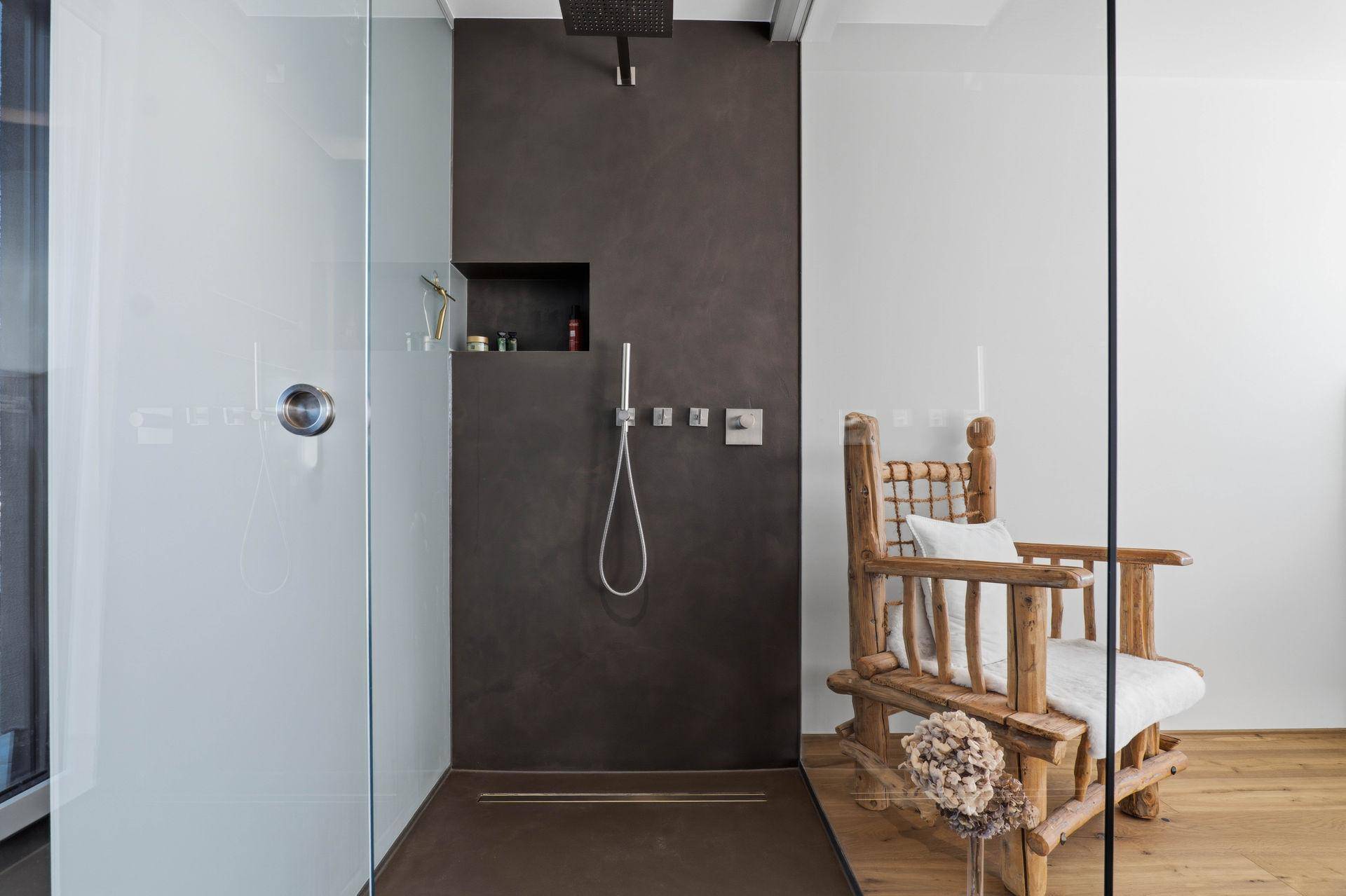 Moderne begehbare Dusche mit dunklen Fliesen und Holzsessel im Badezimmer.