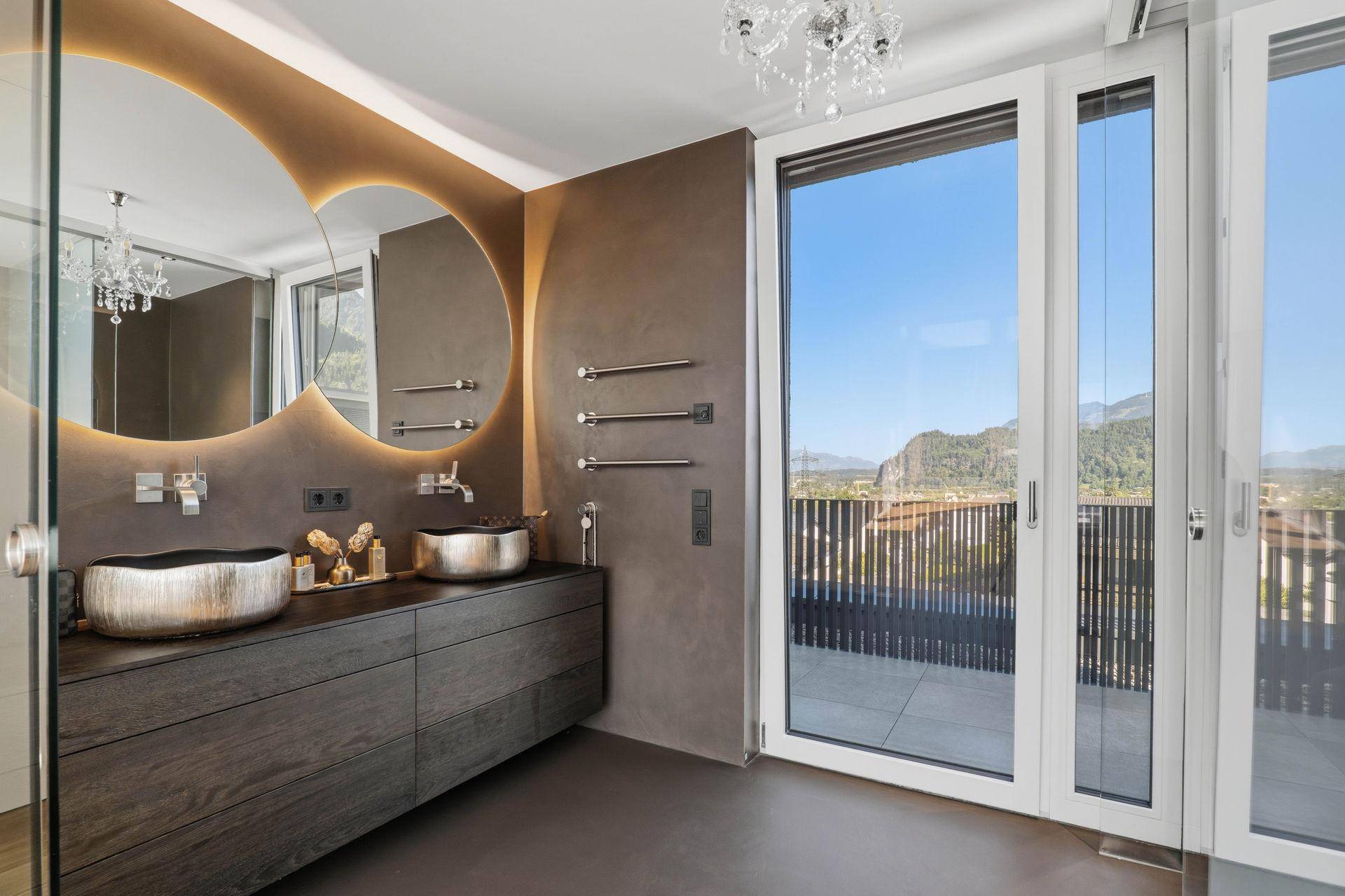 Modernes Badezimmer mit Doppelwaschbecken, runden Spiegeln und Zugang zur Terrasse mit Bergblick.