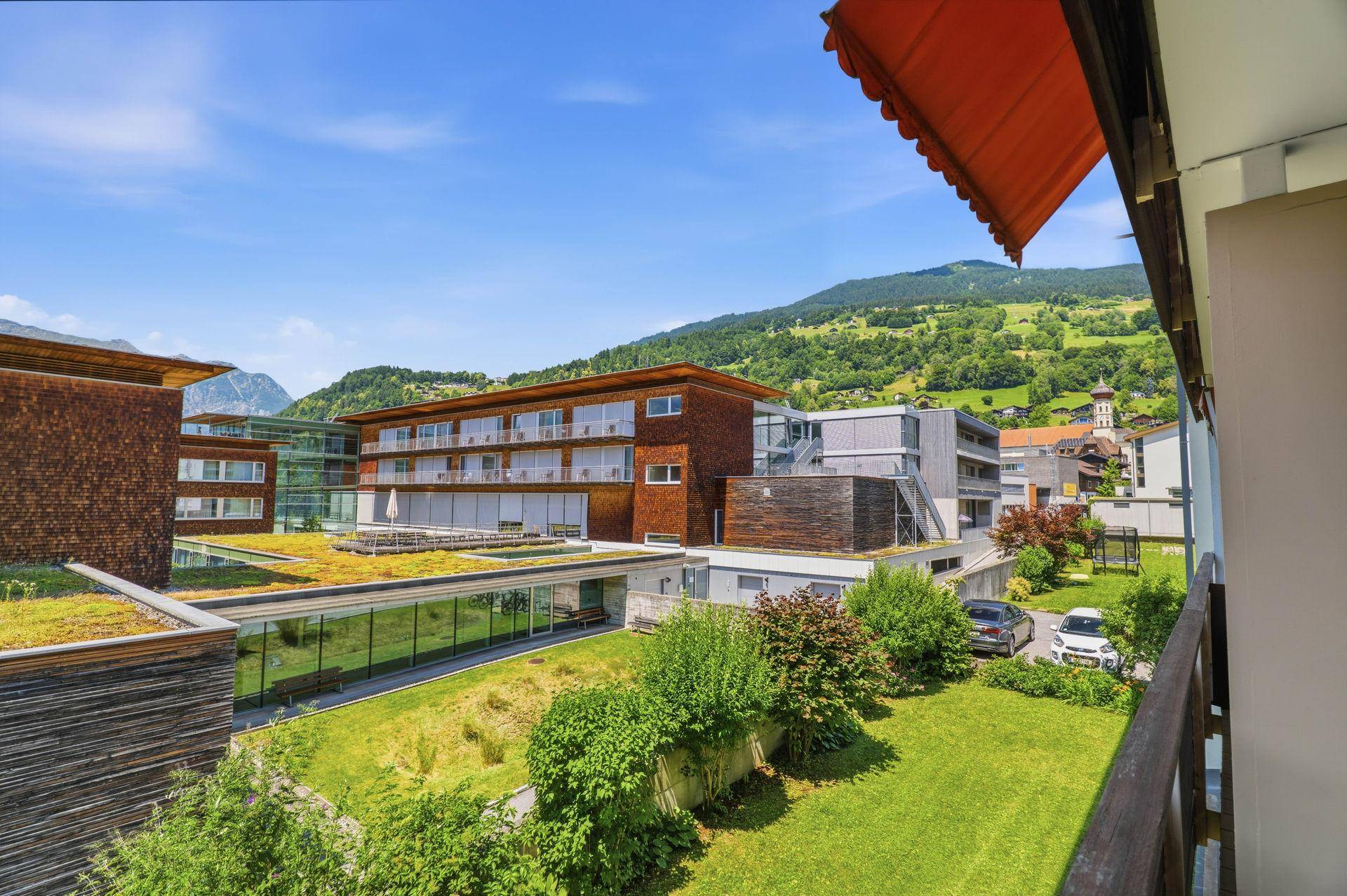 Balkon mit Blick auf moderne Gebäude, grüne Dächer und die umliegende Berglandschaft.