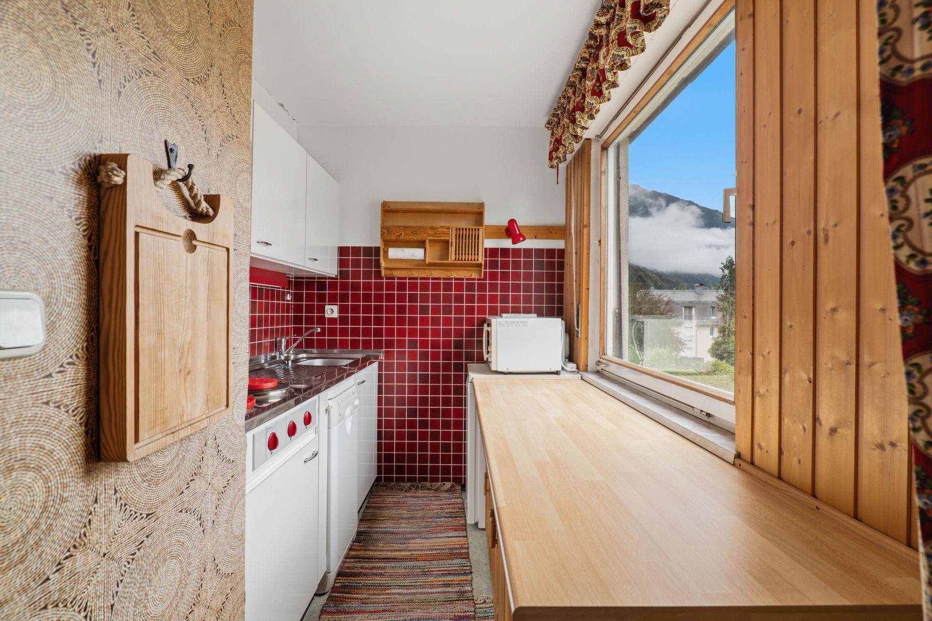 Kleine Küche mit roten Fliesen, Holztheke und Bergblick durch das Fenster.