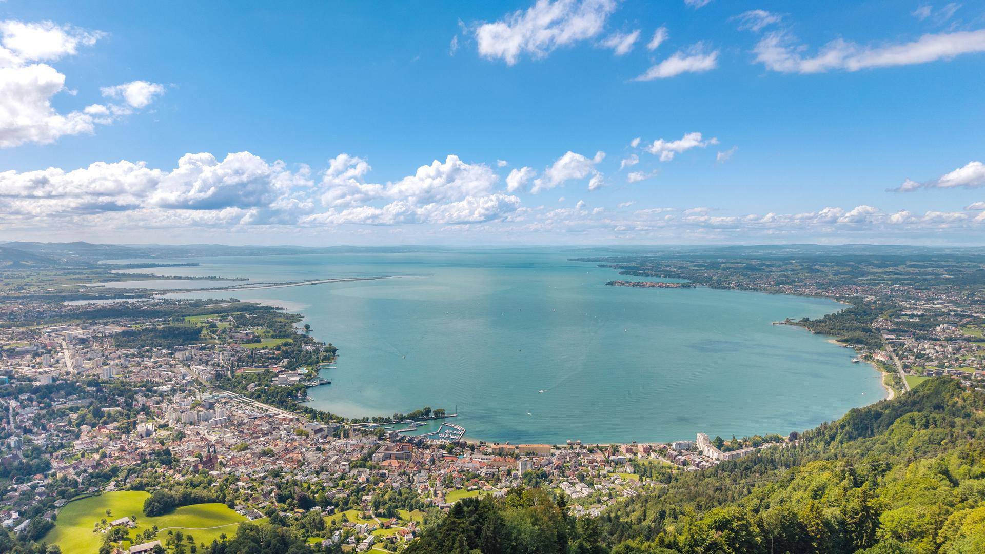 Panoramablick über eine Stadt am See, umgeben von grünen Hügeln unter blauem Himmel.