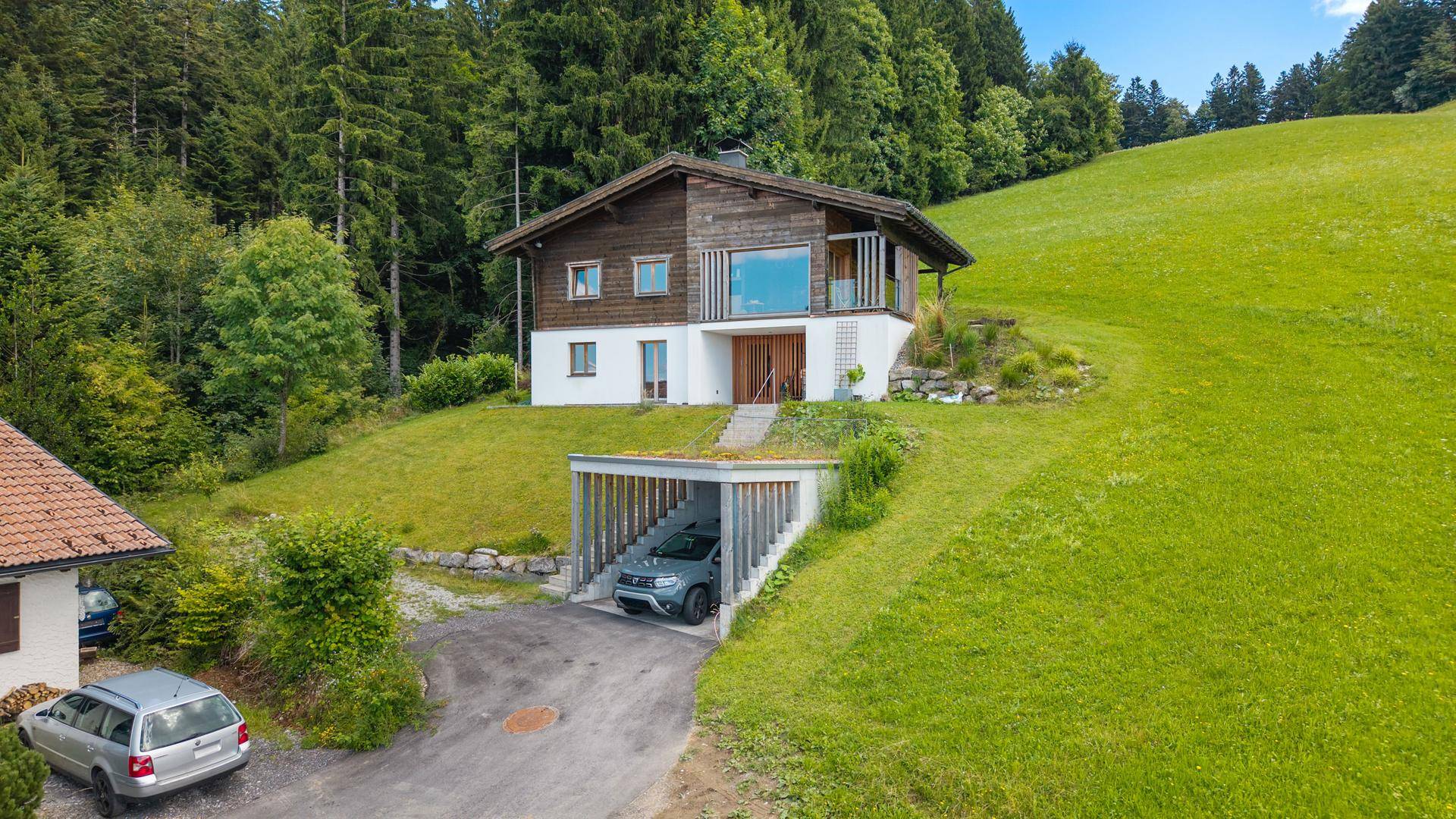 Modernes Haus in Hanglage mit Carport und grüner Umgebung, umgeben von Wald.