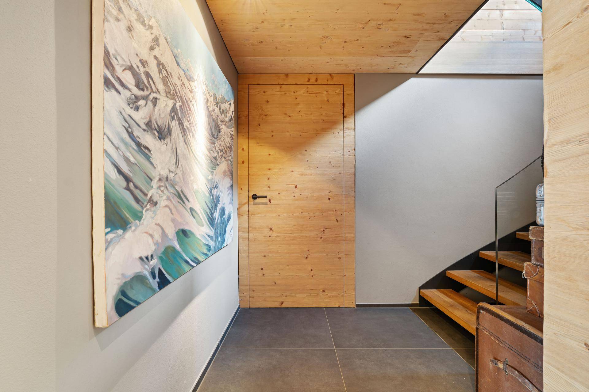 Heller Flur mit Holztür, Kunstwerk an der Wand und einer modernen Holztreppe mit Glasgeländer.