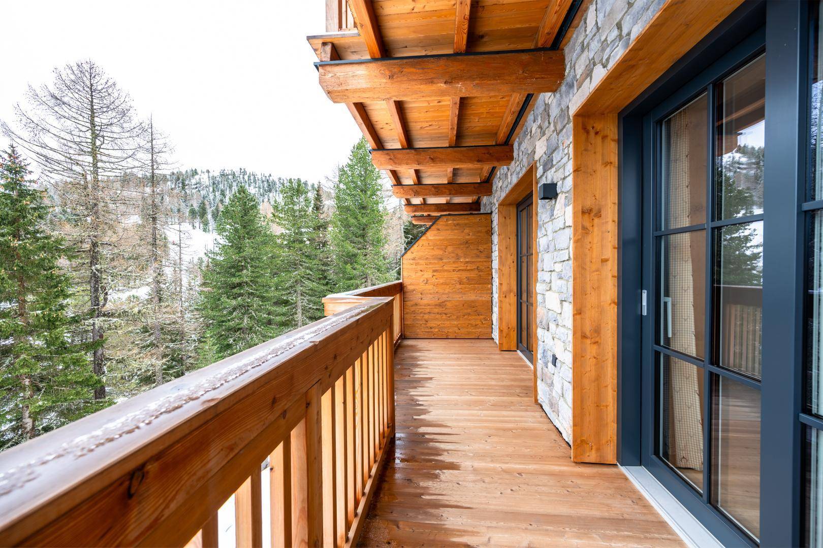 Langer Holzbalkon mit Blick auf die verschneite Berglandschaft und Tannenbäume.