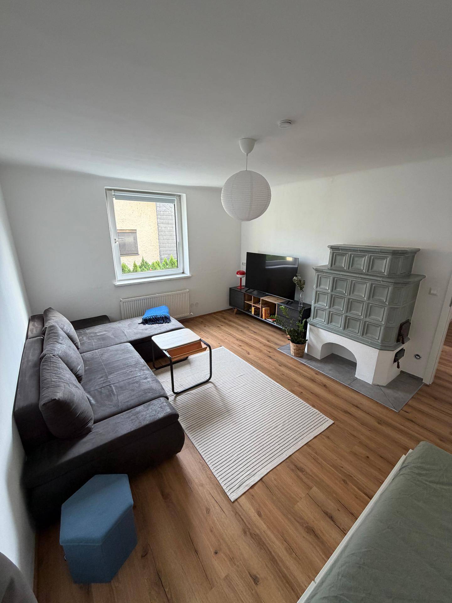 Gemütliches Wohnzimmer mit grauem Sofa, Fernseher und traditionellem Kachelofen.