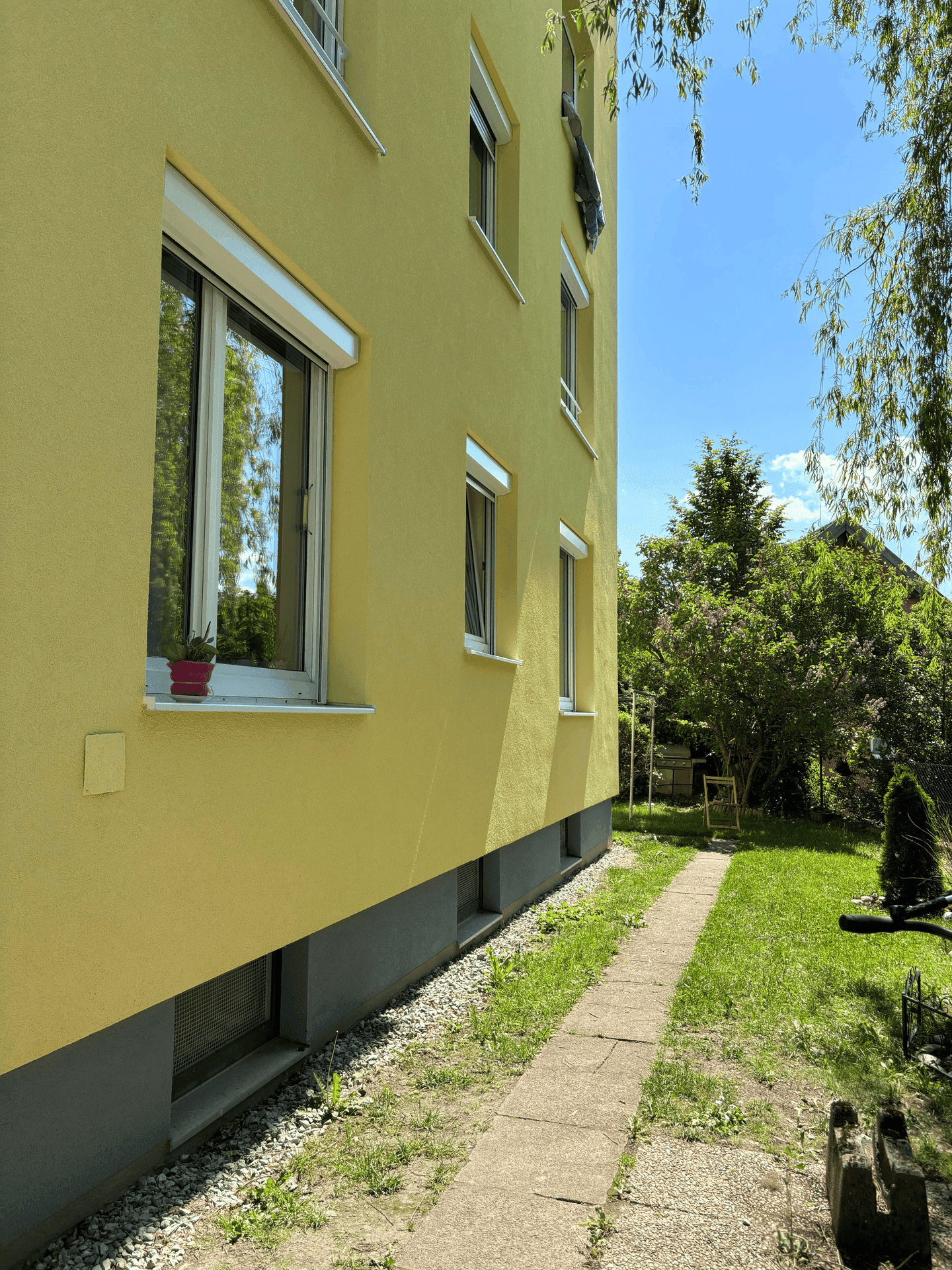 Gelbe Gebäudefassade mit Fenstern und einem Weg, der zu einem grünen Gartenbereich führt.
