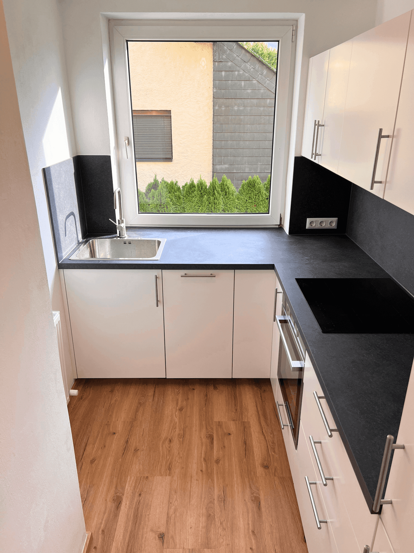 Schmale, moderne Küche mit weißen Schränken, schwarzer Arbeitsplatte und Fensterblick ins Grüne.