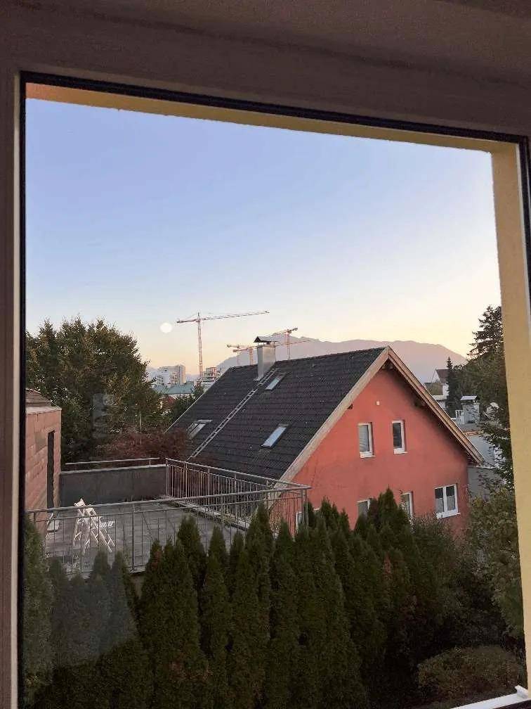 Ausblick aus dem Fenster auf eine Wohngegend mit Häusern, Bäumen und Bergen im Hintergrund.