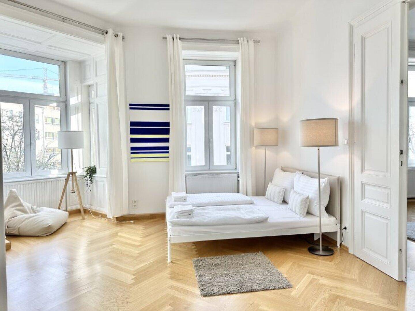 Schlafzimmer mit weißem Bett, zwei Stehlampen und großen Fenstern, die viel Licht hereinlassen.