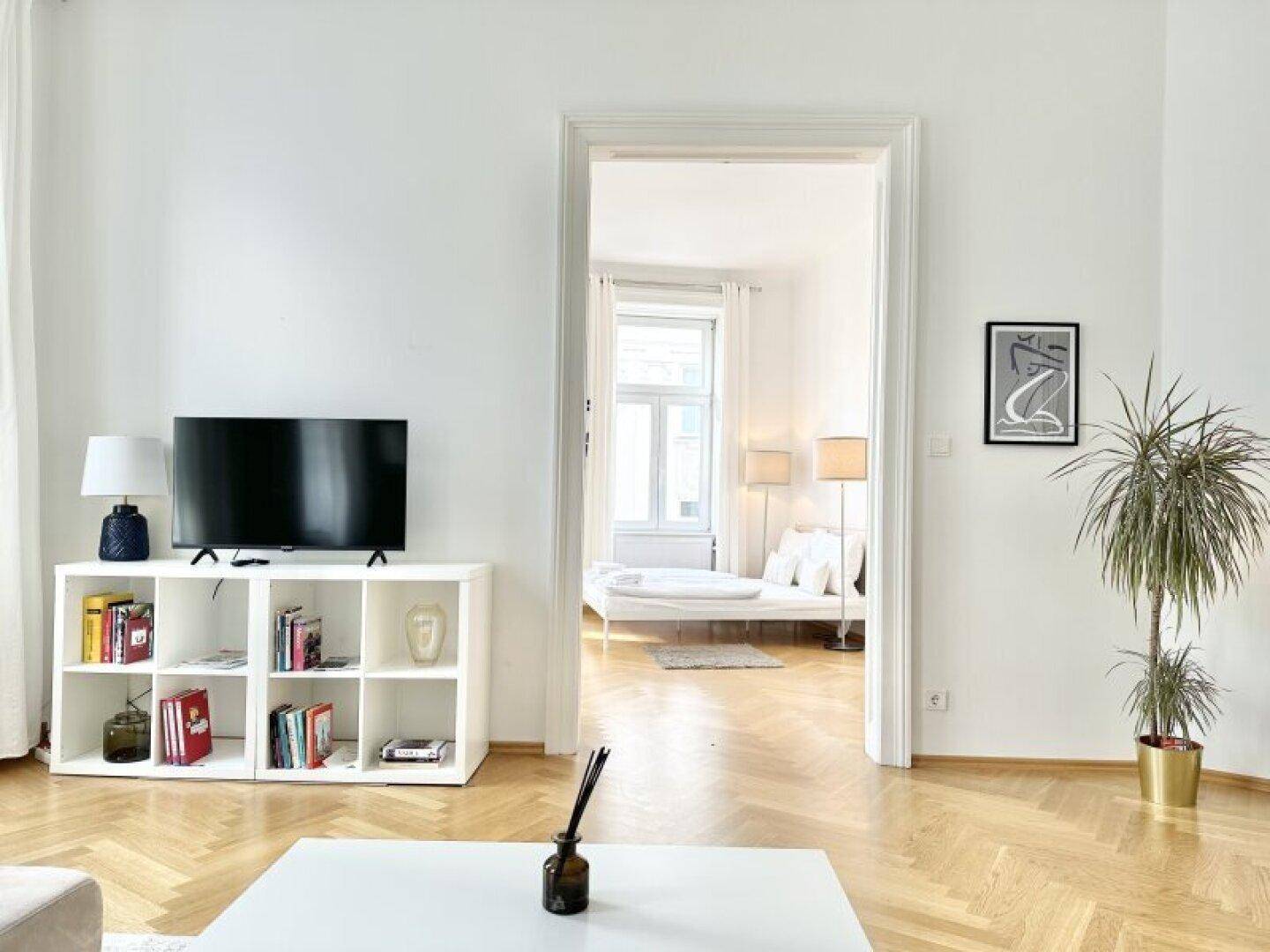 Helles Wohnzimmer mit Fernseher und Blick durch eine offene Tür in ein weiteres Zimmer.