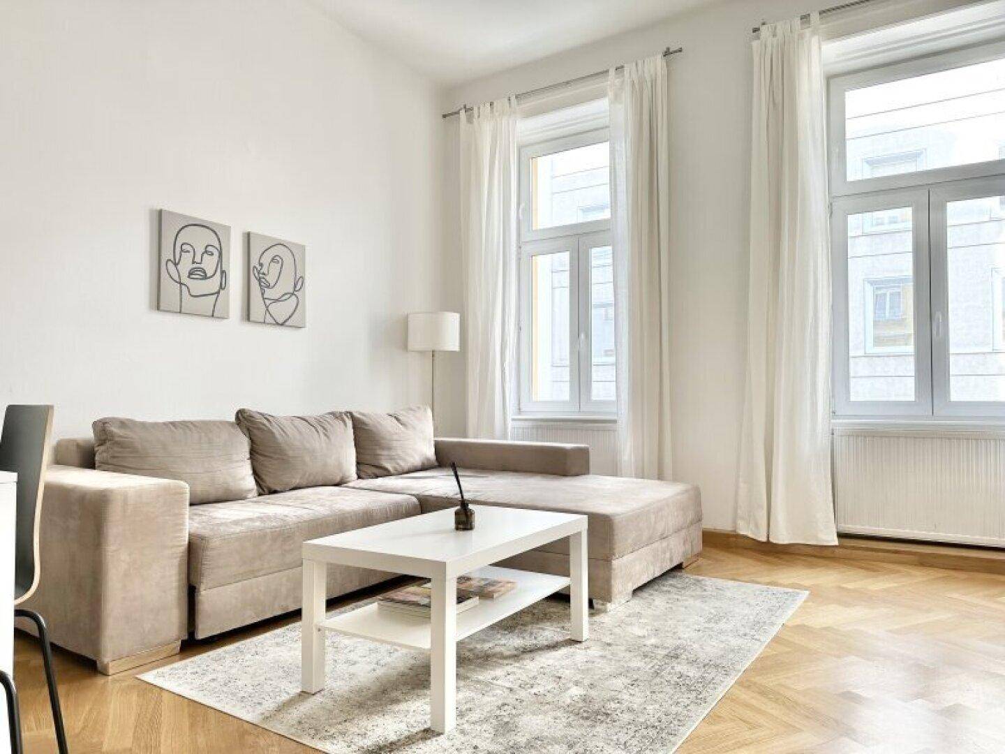 Gemütliches Wohnzimmer mit beigem Ecksofa, hellem Fischgrätparkett und großen Fenstern.