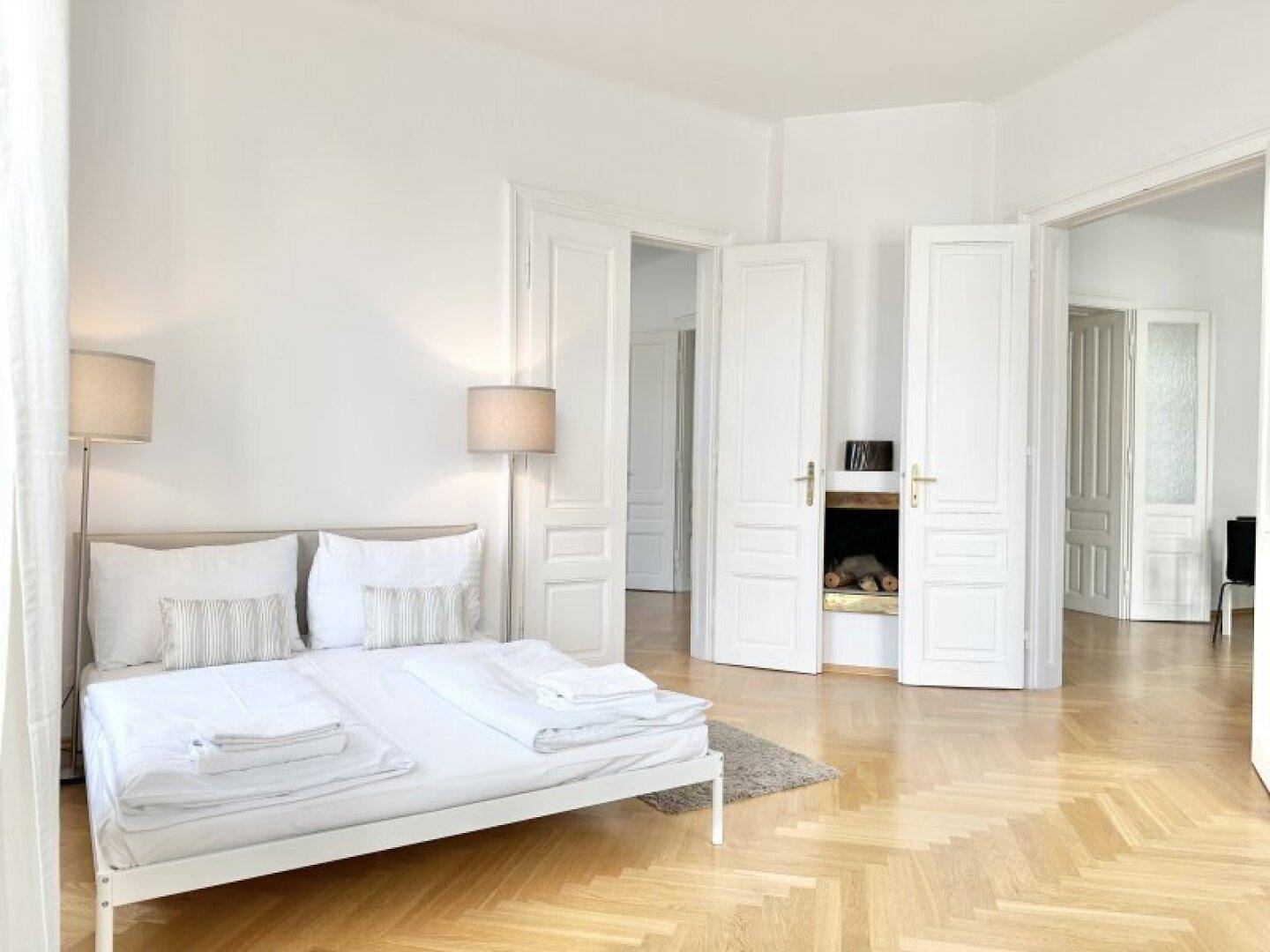 Schlafzimmer mit weißem Bett, Fischgrätparkett und einem dekorativen Kamin in der Wand.