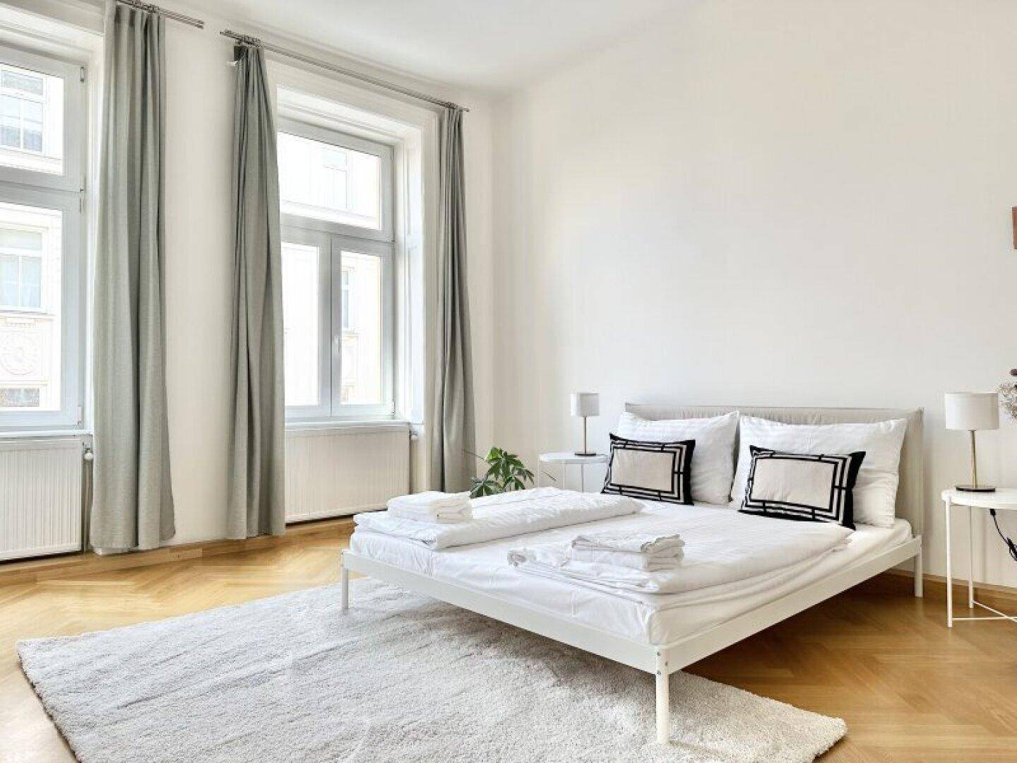 Helles Schlafzimmer mit weißem Bett, Teppich und großen Fenstern mit grauen Vorhängen.