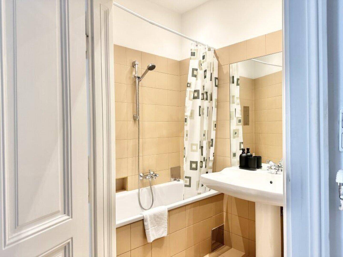 Badezimmer mit Badewanne, Duschvorhang, Standwaschbecken und beigefarbenen Fliesen.