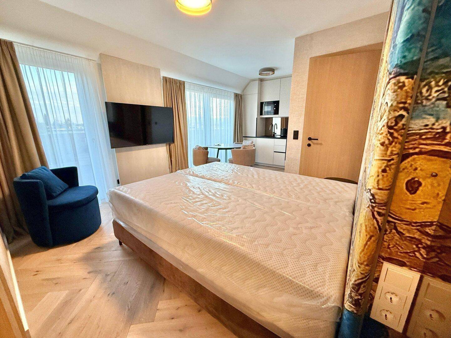 Schlafzimmer mit Doppelbett, Fernseher und kleiner Sitzecke, im Hintergrund eine Küchenzeile.