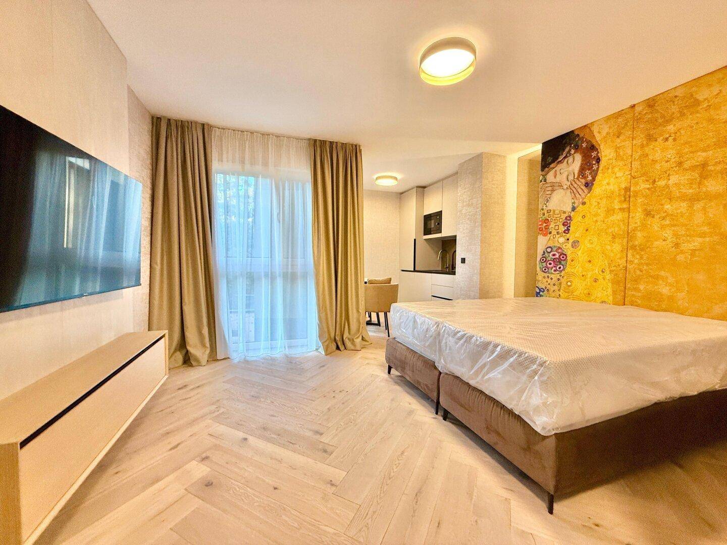 Geräumiges Schlafzimmer mit großem Bett, Wandfernseher und Blick auf die integrierte Küchenzeile.