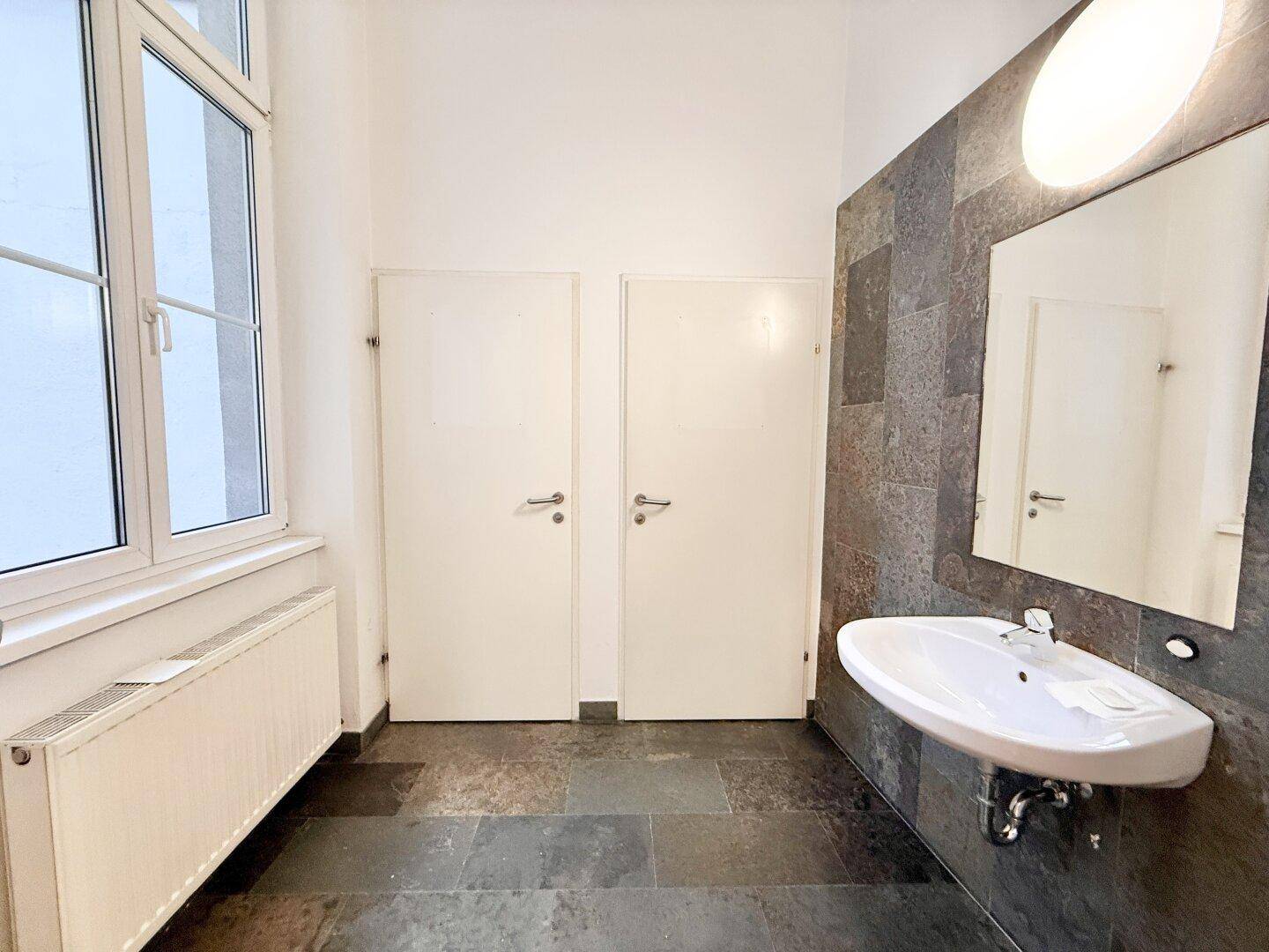 Waschbereich mit modernem Waschbecken, Spiegel und dunklen Fliesen an Wand und Boden.