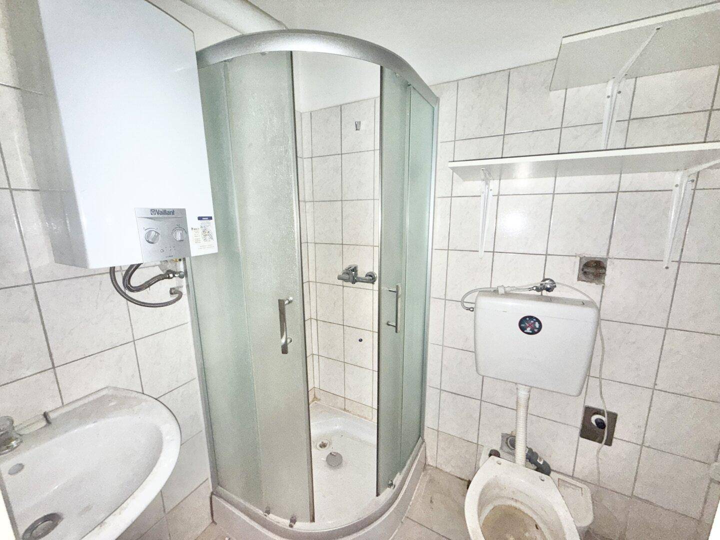 Älteres Badezimmer mit Duschkabine, WC und Boiler, das eine Renovierung benötigt.