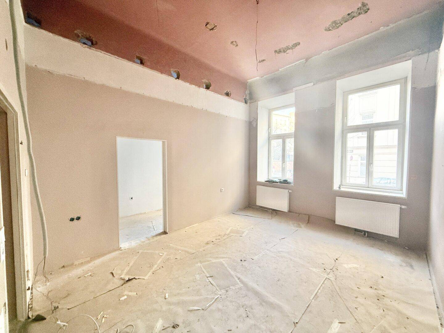Unfertiger Raum mit hohen Decken und einem Durchgang zu einem weiteren Zimmer.