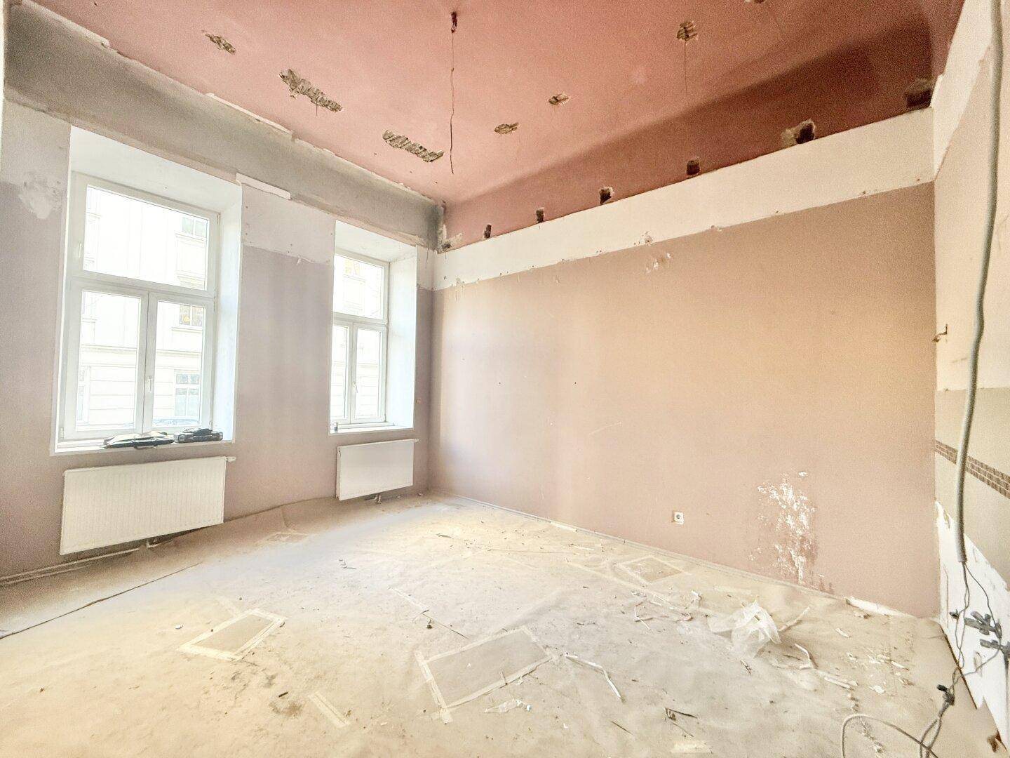 Renovierungsbedürftiger Raum mit hohen Decken, zwei Fenstern und vorbereiteten Wänden.