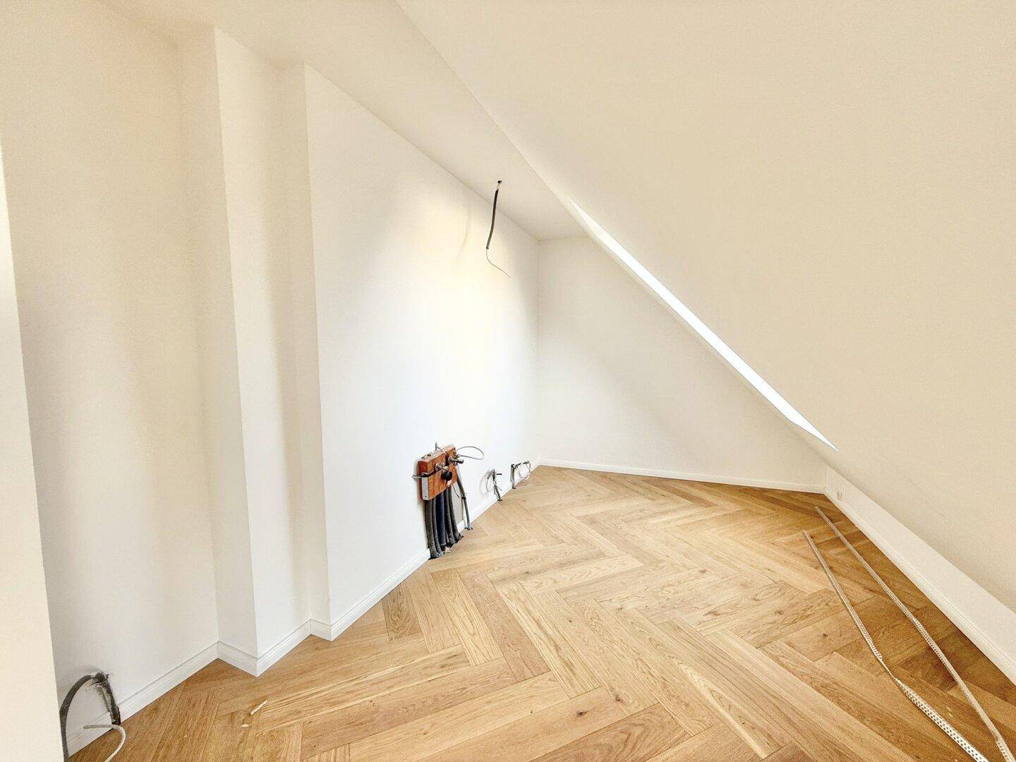 Leerer Raum mit Dachschräge, hellem Parkettboden und sichtbaren Elektroinstallationen.