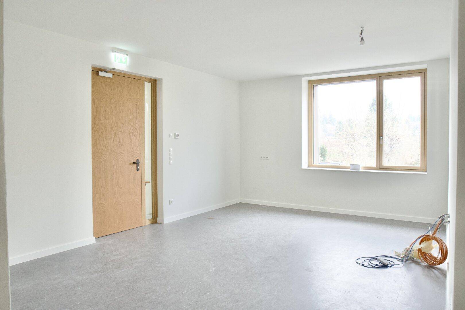 Geräumiger, leerer Raum mit Holztür und großem Fenster, das viel natürliches Licht hereinlässt.