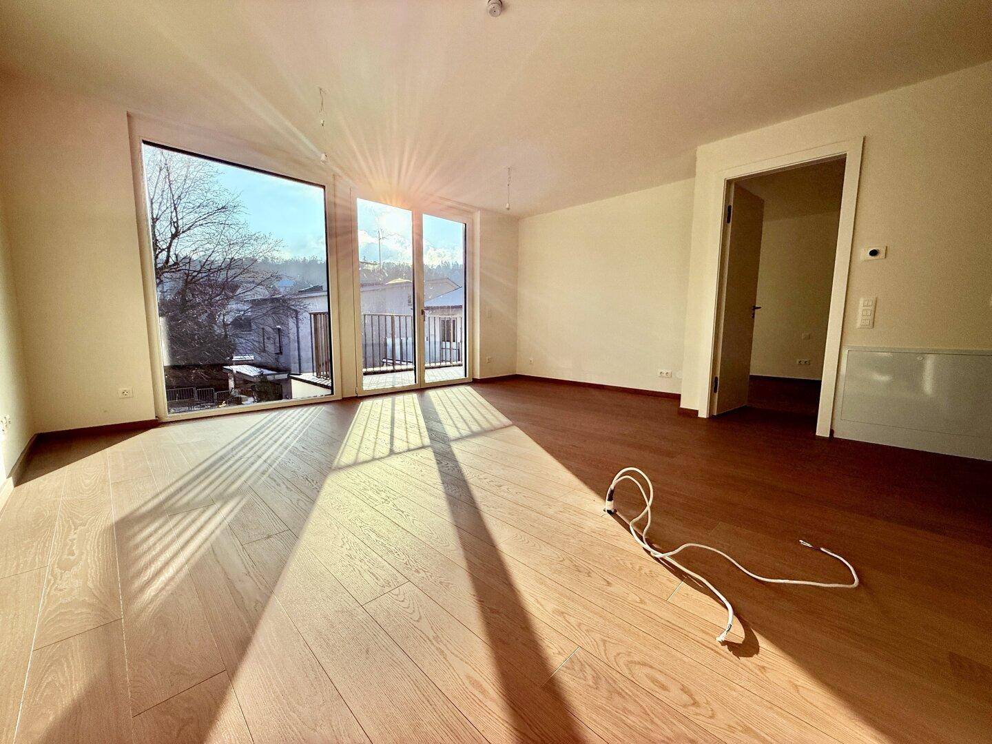 Heller, leerer Wohnbereich mit großen Fenstern, Holzboden und Zugang zu einem Balkon.