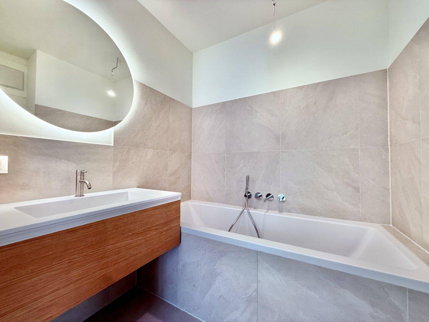Modernes Badezimmer mit Badewanne, Waschtisch und beleuchtetem Spiegel, hell gefliest.