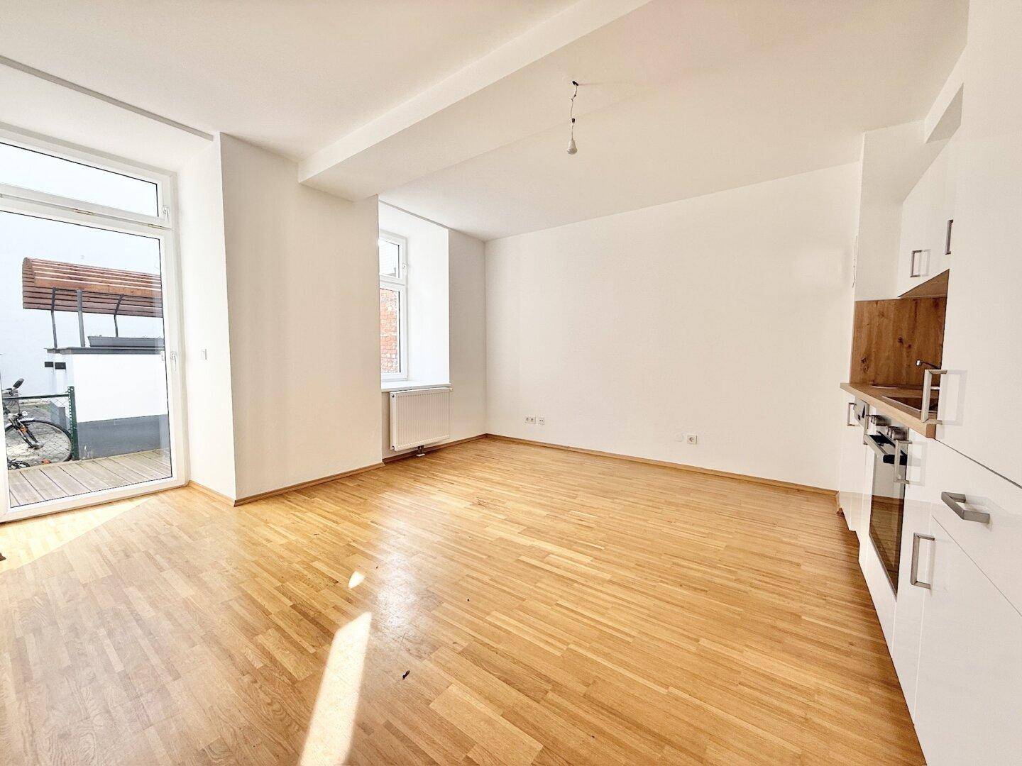 Leerer Wohnbereich mit Holzboden, vorbereiteter Küchenzeile und direktem Zugang zur Terrasse.