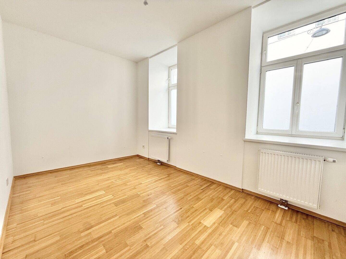 Heller, leerer Raum mit zwei Fenstern und Holzboden, ideal für verschiedene Nutzungsmöglichkeiten.