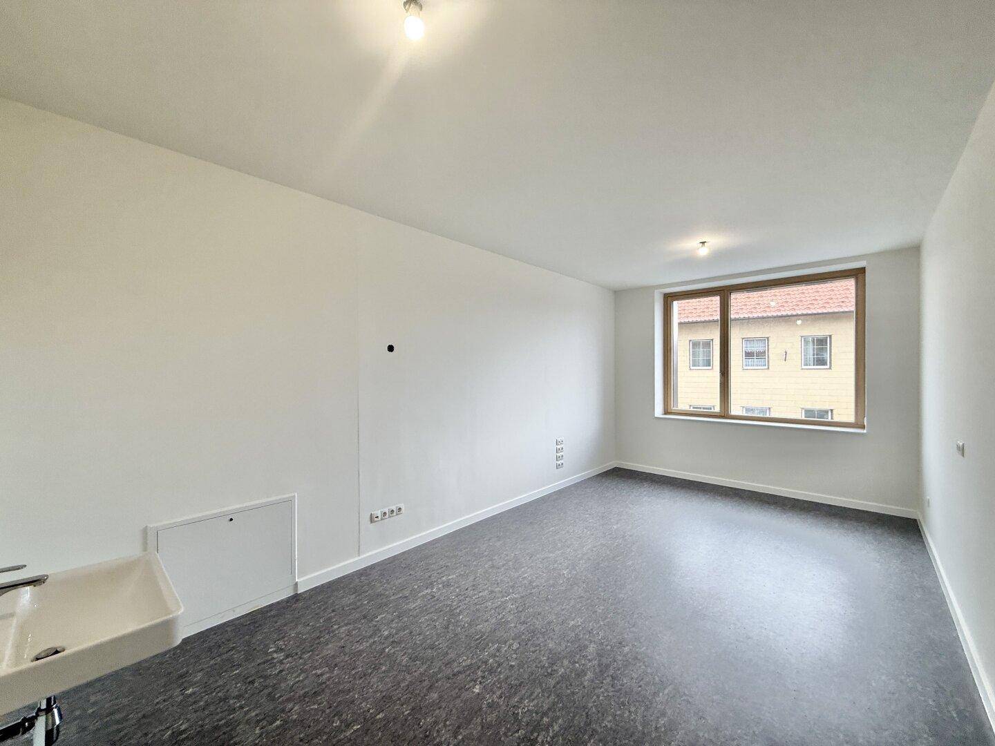 Leerer Raum mit Waschbecken, einem Fenster und dunklem Boden, ideal für eine Praxis.