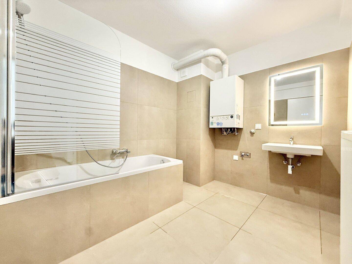 Modernes Badezimmer mit Badewanne, Duschabtrennung, beleuchtetem Spiegel und beigefarbenen Fliesen.