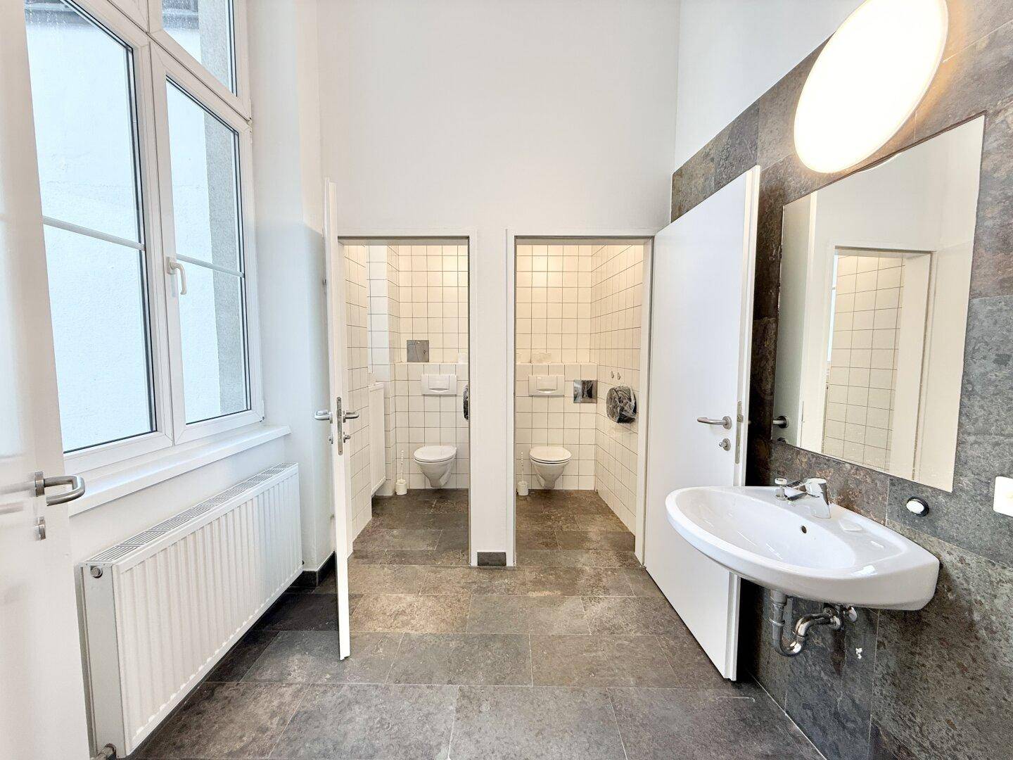 Sanitärbereich mit Waschbecken, Spiegel und zwei separaten Toilettenkabinen.