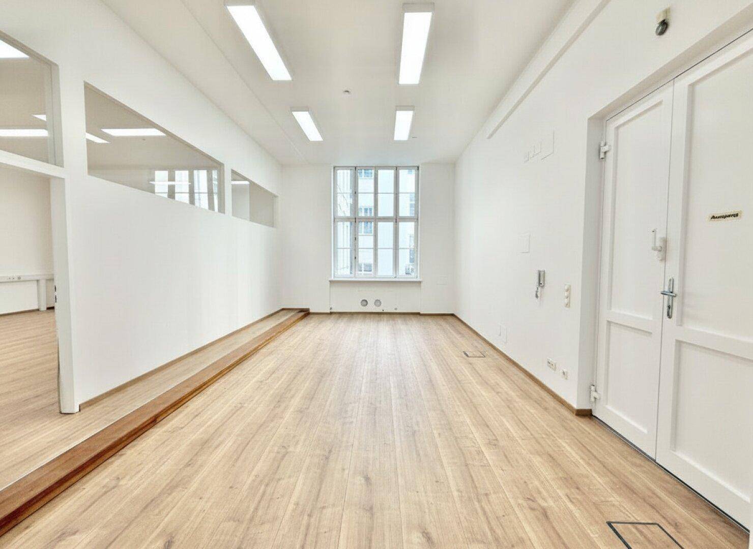 Leerer Büroraum mit Holzboden, weißen Wänden und modernen Deckenleuchten.