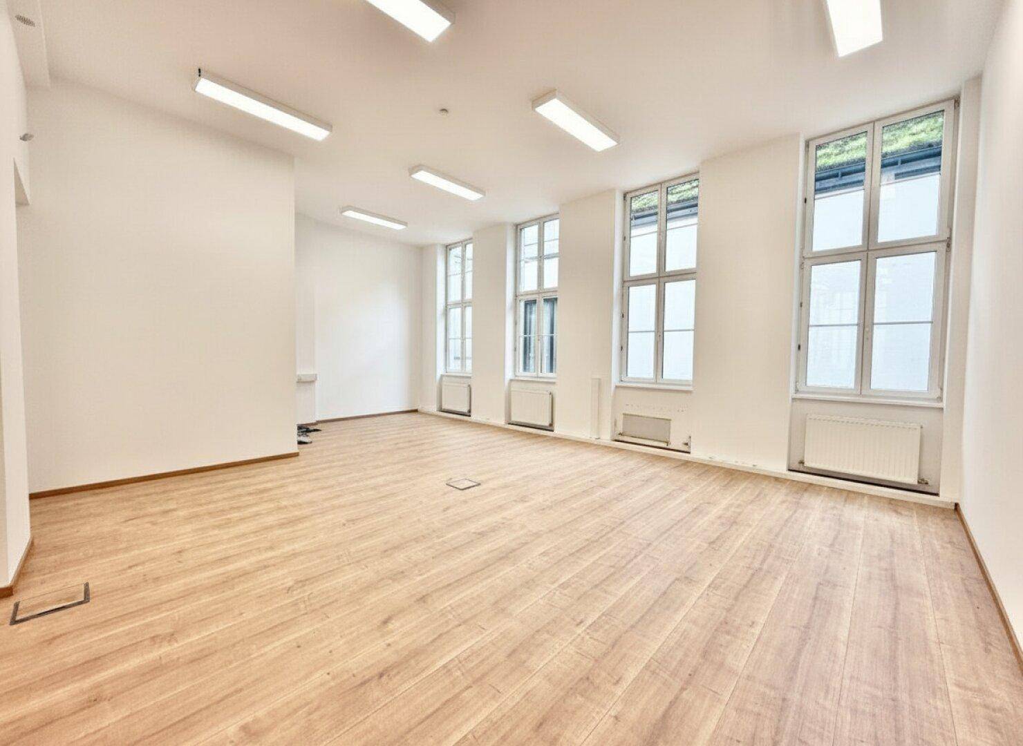 Heller, leerer Büroraum mit mehreren hohen Fenstern und Holzboden.