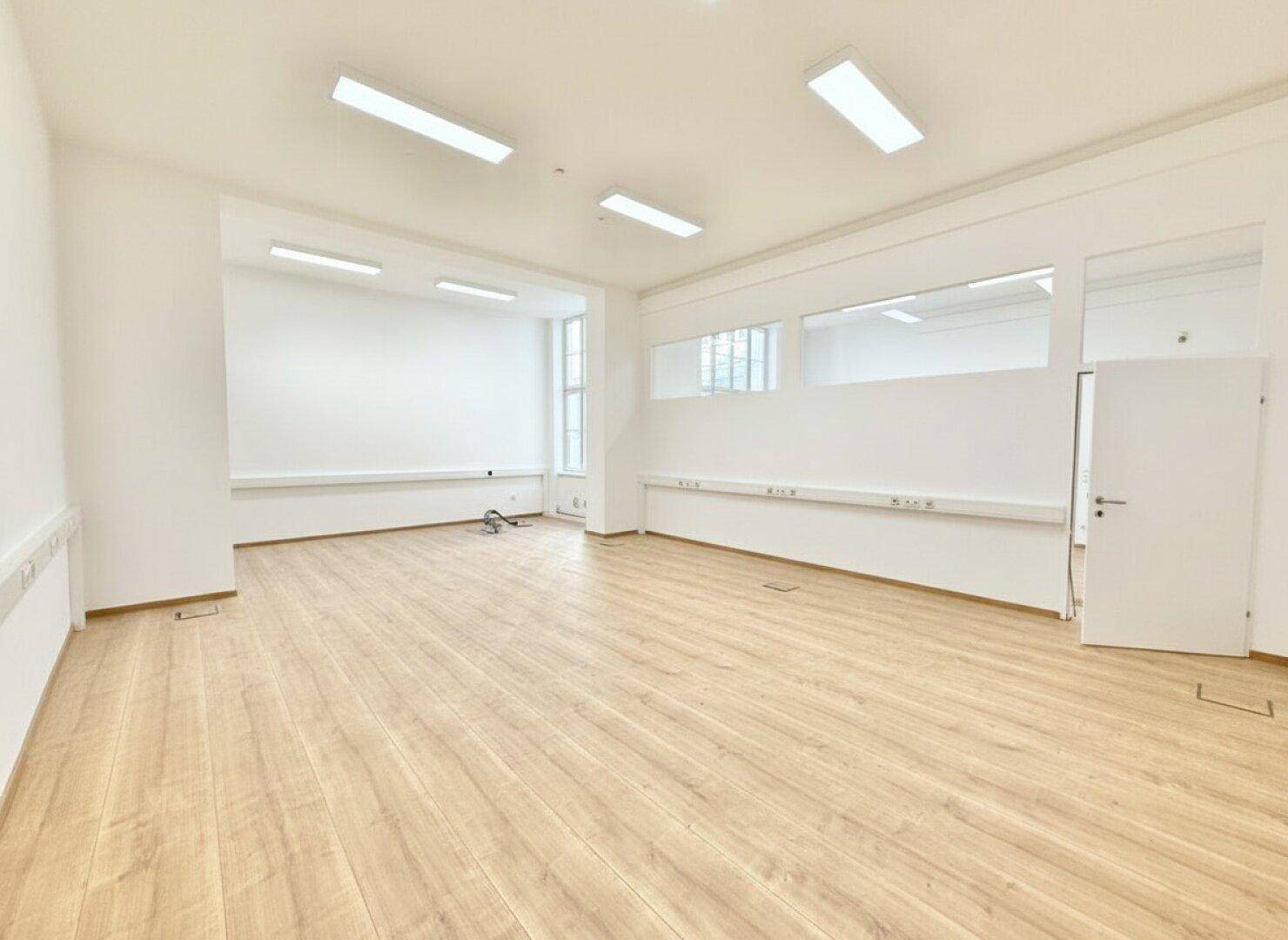 Geräumiger, leerer Bürobereich mit Holzboden und Trennwänden mit Glaselementen.