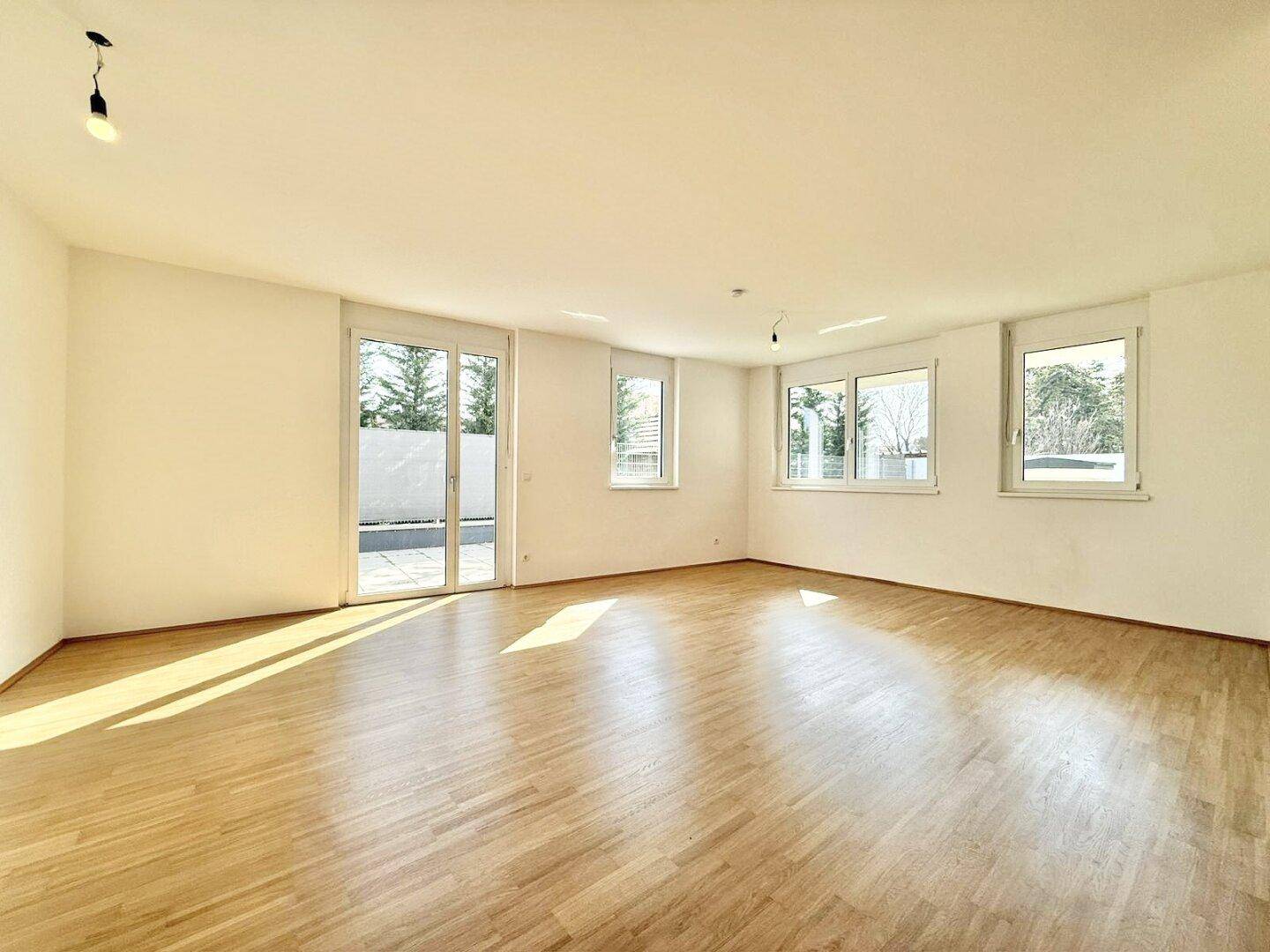 Heller, leerer Wohnbereich mit Holzboden, großen Fenstern und viel natürlichem Licht.