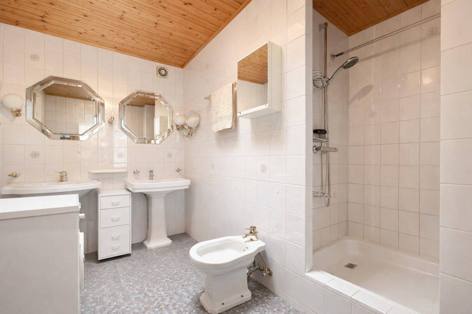 Helles Badezimmer mit zwei Waschbecken, Dusche, Bidet und Holzdecke.