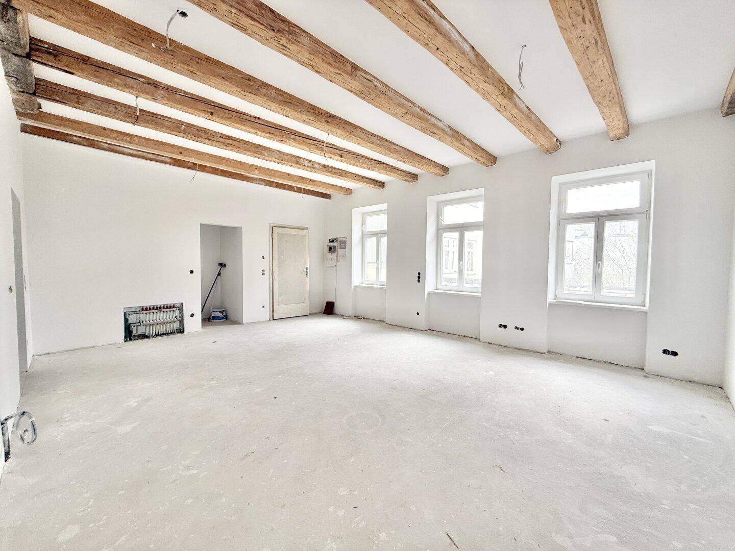 Großer, leerer Raum mit freiliegenden Holzbalken und mehreren Fenstern, im Rohbauzustand.
